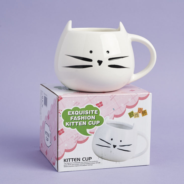 a white mug shaped like a cat's head, complete with black whiskers, nose, and eyes, sitting on top of its box