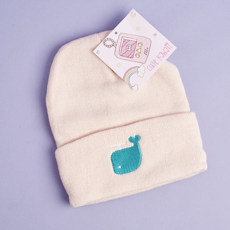 a pale pink beanie hat featuring a turquoise whale embroidered on it with its tag