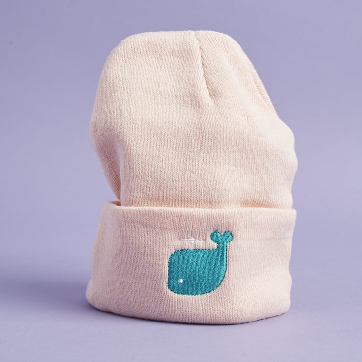 the front of a pale pink beanie hat with a turquoise blue cartoon whale embroidered on the front